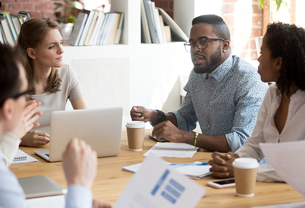 Group of peers sit at table and discuss over coffee and computers