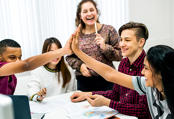 Students reach across table to high five and teacher smiles overlooking them