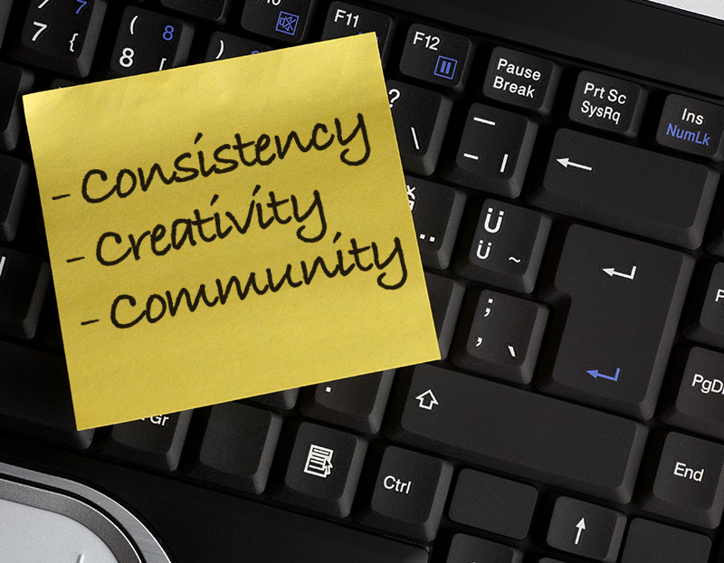 Computer keyboard has sticky note in it with words, "consistency, creativity, and community" written on it