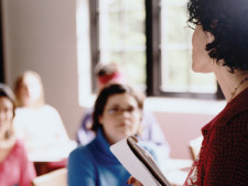 Student in background observes teacher holding papers in front of classroom