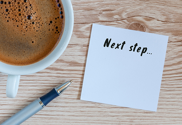 Coffee and pen sit on table with a notepad that says, "Next step..."