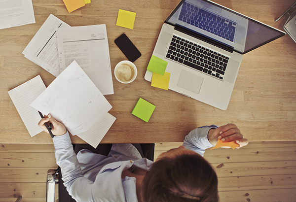 Person sits at desk with papers, stickies and coffee sprawled out with computer opened