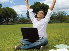 Person sits outside with computer on lap and stretches arms into the air with excitement