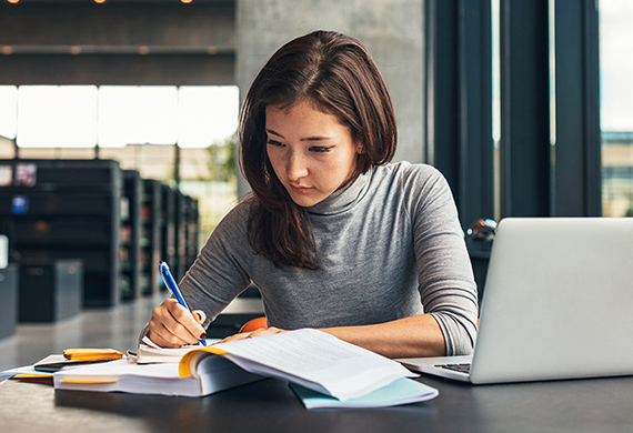 Woman looks at notes and writes in notebook with computer screen opened