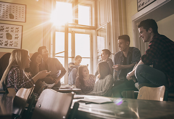 Students sit and stand over table while talking and sun shines through window