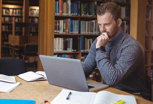 Man sits at desk while looking at computer screen with library books in background