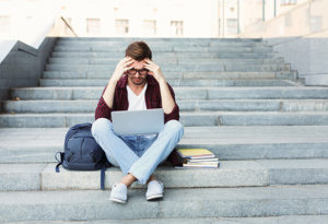 Student sits on stairs with head in hands looking at laptop
