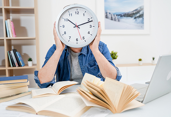 Person holds up clock in replace of their head with books and computer laid out in front of them